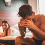 Man sitter i sängen med huvudet i händerna. Kvinna i bakgrunden,