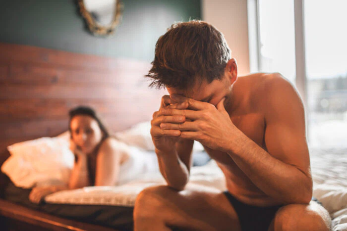 Man sitter i sängen med huvudet i händerna. Kvinna i bakgrunden,